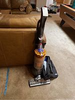 Dyson upright vacuum cleaner with clear yellow dust container, hose attachment, and black accessory bag shown in living room