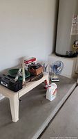 Photo showing plastic patio table with various items including black tool case, green garden hose coil, cordless drill, Orby laser leveler, insect killer spray, small basket, and part of a fan base.