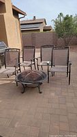 Four metal patio chairs with tan mesh seats and backs arranged in a patio setting with the fire pit placed in front of them on a paved surface.
