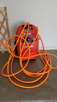 Front view of red Husky Air Scout air compressor with connected long orange air hose coiled on the floor.