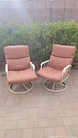 Two outdoor swivel rocking patio chairs with reddish-brown cushions and beige/off-white metal frames, placed on a patio with green bush background.