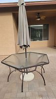 Full view of hexagonal glass top patio table with closed crank-up umbrella and metal frame on paved patio surface.