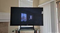 Front view of a large 50 inch plasma TV powered off, black frame, mounted on a glass stand reflecting room interior.