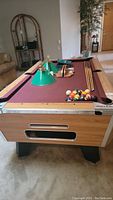 Full view of pub/tavern style pool table with maroon felt, wooden rails, four cues and ball set on top.