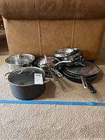 Full view of the collection of pots, pans, and lids displayed on carpeted floor in front of a tan sofa