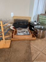 Overall view showing roaster pan, stock pot, Crock-Pot, flatware in wooden trays, wooden cup rack, and glassware in background.