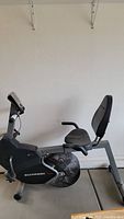 Side view of the Schwinn stationary recumbent bike showing the padded seat with backrest, large fan wheel, and digital monitor mounted on the frame.