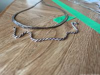 Close-up of two silver chains, one twisted rope style and one flat snake chain laying on a wooden table.