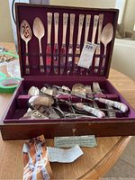 Full view of the cutlery set in wooden box showing knives, forks, spoons, and serving utensils inside burgundy lined compartments with labels and info cards.