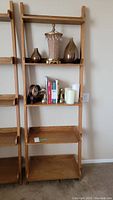 Front view of solid wood leaning shelf unit with items on four shelves including decorative vases, books, candles, and a clock. The bottom shelf is empty.