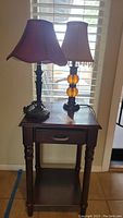 Photo of wood grain accent table with drawer and lower shelf, holding two lamps: one amber glass orb style with fringe shade, one metal with red bell-shaped shade