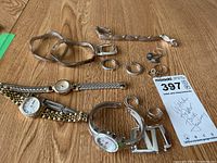 Full view of the three watches laid flat with assorted silver toned jewelry including several bracelets, earrings, and rings on wooden surface