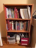 Front view of light brown wood 3-shelf book shelf. Three shelves holding books and two plastic storage bins labeled 'Shoe Accessories'. Shelf is rectangular and in good condition.
