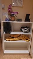 Front view of white book shelf with 4 shelves, some miscellaneous items on shelves including a flower pot, picture frame, tissue box, jacket, helmet, and other small items; shelf dimensions approximately 31.5x11x42 inches. Shows scuffs and surface wear.