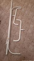 Disassembled white metal clothing rack parts laid on carpeted floor showing vertical support bars and curved braces.