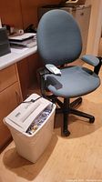 Steelcase office chair with Fellowes paper shredder beside it, showing front side of chair with armrests and blue upholstery.