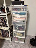 Front view of the white plastic 7 drawer storage cabinet containing various craft supplies inside clear drawers, positioned next to a white shelf with records.