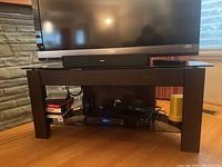 Front view of the wood and glass TV stand with TV and soundbar on top, electronic devices on glass shelf below
