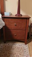 Front view of one wooden nightstand showing two drawers with metal knobs, a red lamp on top, and a patterned rug below. Visible wear and scratches on the wood.
