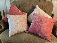Four cushions, two pink and two blush, arranged on a patterned sofa. Pink cushions have a diagonal pleated velvet-like design. Blush cushions have a geometric quilted texture.
