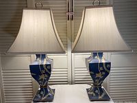 Pair of blue lamps with hand-painted gold floral vine designs and white fabric trapezoidal lampshades.