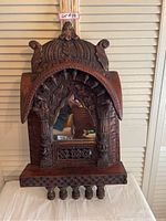 Front view of ornate wooden mirror with detailed carvings and central mirror glass, set on a white cloth against a louvered backdrop.