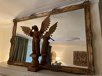 Full view of the large ornate mirror with detailed frame and a wooden angel figurine reflected in it.