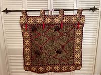 Full view of tapestry hanging on rod against white louvered background showing design, tassels, and loops.