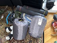 Photo showing 2 sleeping bags in gray stuff sacks, 2 rolled yoga mats (black and gray), and partial view of a dark backpack on a patterned carpet.