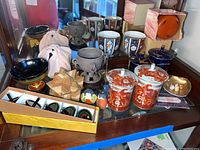 Photo showing entire lot on glass table including enamel goblets, wooden puzzle, spinning tops in box, porcelain containers, and various bowls.