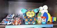 Photo showing a Furby plush toy, three LEGO vehicles, and several wooden rattle toys on a dark shelf