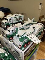 Photo of three Hess toy trucks including a large truck with airplane attachment on top and two smaller trucks in green, white, and red color scheme, placed on their original boxes.