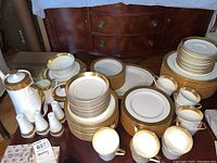 Full view of Mikasa bone china dinnerware service showing stacked plates, cups, and serving pieces with gold trim.