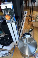 Photo of large stainless steel pot with lid, two non-stick fry pans, and boxed rotisserie kit against a wooden floor background.