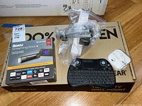 Roku Streaming Stick+ retail box, wall mount box, mini keyboard, earbuds, and mounting hardware in plastic bags on wooden floor