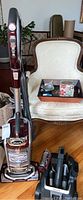 Full view of Shark upright vacuum in burgundy with attachments in caddy next to it on floor and near beige chair