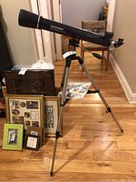 Full view of the Celestron AstroMaster LT 70 telescope mounted on a tripod with lenses and accessories on the floor nearby.