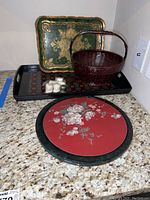 Green and gold rectangular lacquer tray, black lacquer tray, wicker basket, round floral stone plate, and small white items on countertop