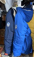Two winter coats, dark blue and bright blue, hanging side by side showing coat patches and outer shell.
