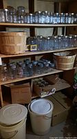 Four wooden shelves filled with rows of clear glass canning jars with metal lids, two wooden bushel baskets on shelves, white and beige plastic pails with lids on the floor, and boxes below shelves.