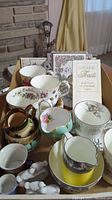 Various tea cups, saucers, creamers and small decorative items including a wedding toasts book, doves napkin rings and a 25th anniversary trivet arranged on a table.