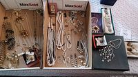 Top-down view of two boxes containing assorted costume jewelry including necklaces, chains, earrings, brooches, and persona charm boxes.