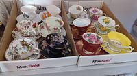 Photo showing twelve assorted porcelain cups and matching saucers with varied floral and decorative patterns in two boxes.