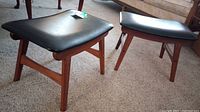 Two black vinyl padded foot stools with teak legs placed side by side