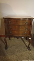 Front view of the Gibbard side table showing three drawers, brass ring pulls, curved cabriole legs, and carved claw feet on carpeting.
