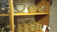 Top shelf displays clear pressed glass candy dish with lid and medium-sized scalloped-edge fruit bowl; lower shelf shows eight filigree-holder glasses in caddie