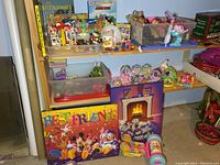Photo showing a shelf with various toys including a plastic container holding a Playmobil merry-go-round ride and several Playmobil figures and accessories, children's storybooks, and Disney dishware items.