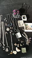 Top-down view of multiple silver-toned necklaces, rings, earrings, brooches, and a grey jewellery pouch with pink decorations, spread on a black surface.