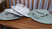 Four hats in two styles and colors on wooden chair, including light beige/gray and green hats with chin straps