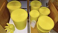 Seven yellow Tupperware items including canisters, round containers, measuring cups, and scoop in kitchen cabinet.